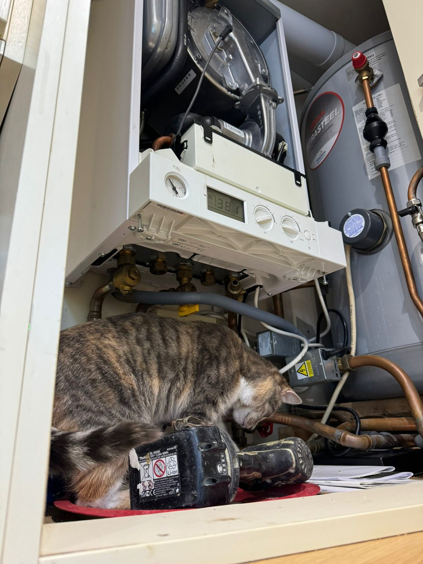 Wall-hung gas boiler with front panel removed showing internals, alongside an unvented cylinder and copper pipework in a domestic airing cupboard