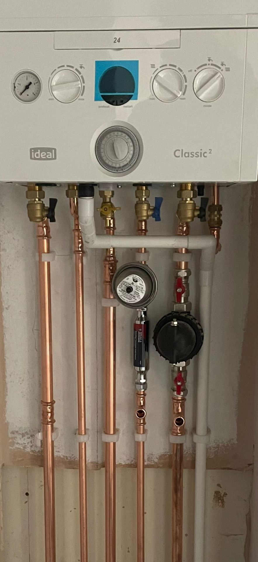 Ideal Classic gas boiler with front panel showing gauges and controls above a manifold of copper pipework, system filter and isolation valves